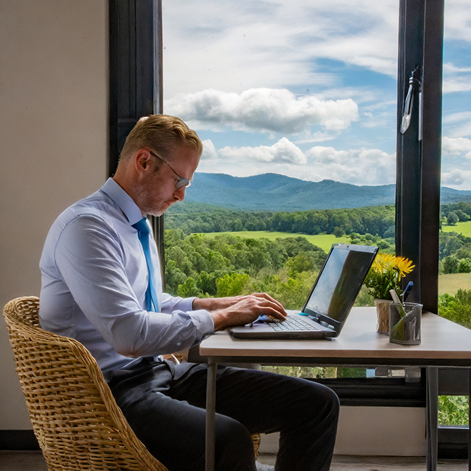 Man in business attire sitting with a laptop in front of a window looking out over fields